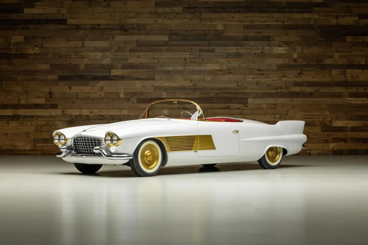 Fehér színű, nyitott Cadillac Elegante roadster aranyozott díszítőelemekkel fa háttér előtt