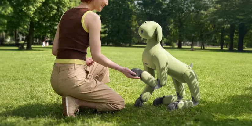 Zöld textilburkolatú robotkutya pacsit ad egy fűben guggoló nőnek a parkban.