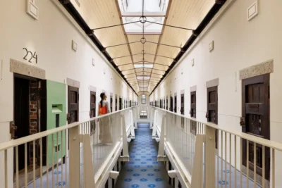 Hosszú, fehér falú börtönfolyosó kék mintás szőnyeggel és faajtókkal